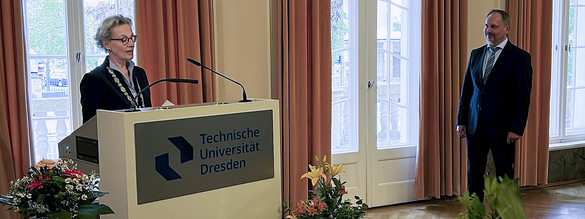 Ceremonial Address: TUD President Staudinger speaks from a lectern in the Rectorate’s banquet hall. Several large bouquets of flowers are arranged around her. Prof. Mechtcherine, the honoree, stands next to the lectern and listens to her.