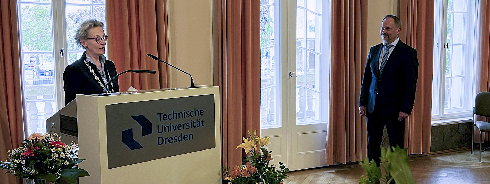 Festrede: TUD-Rektorin Staudinger spricht an einem Pult im Festsaal des Rektorats. Um sie herum stehen mehrere große Blumensträuße. Prof. Mechtcherine steht als zu Ehrender neben dem Rednerpult und hört ihr zu.