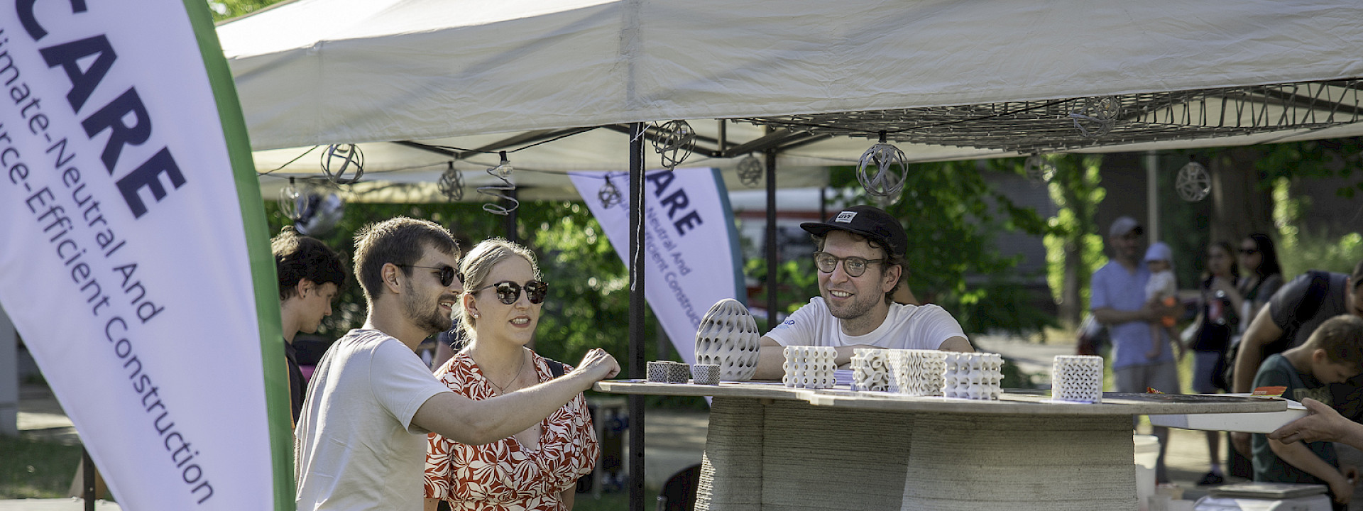 Three people are standing at a high table. Next to them is a banner bearing the name of the CARE Cluster of Excellence. On the table are fist-sized and delicate concrete exhibits, with small information cards next to them. The third person is wearing a CARE T-shirt and is explaining the exhibits to the two visitors.