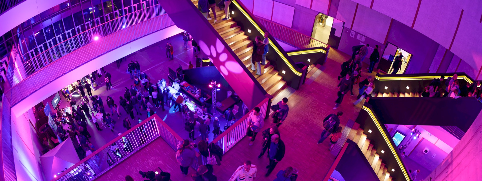 A full house bathed in atmospheric lighting: numerous guests bring the C.A.R.L to life during the RWTH Science Night. A wide-angle shot of a well-attended evening event in the modern, multi-storey foyer of a building. The entire scene is bathed in intense pink and purple light. In the centre, a large, striking wooden staircase leads upwards, with people moving along it. Numerous people are standing and walking on the main floor and the gallery levels; there are information stands and event lighting.