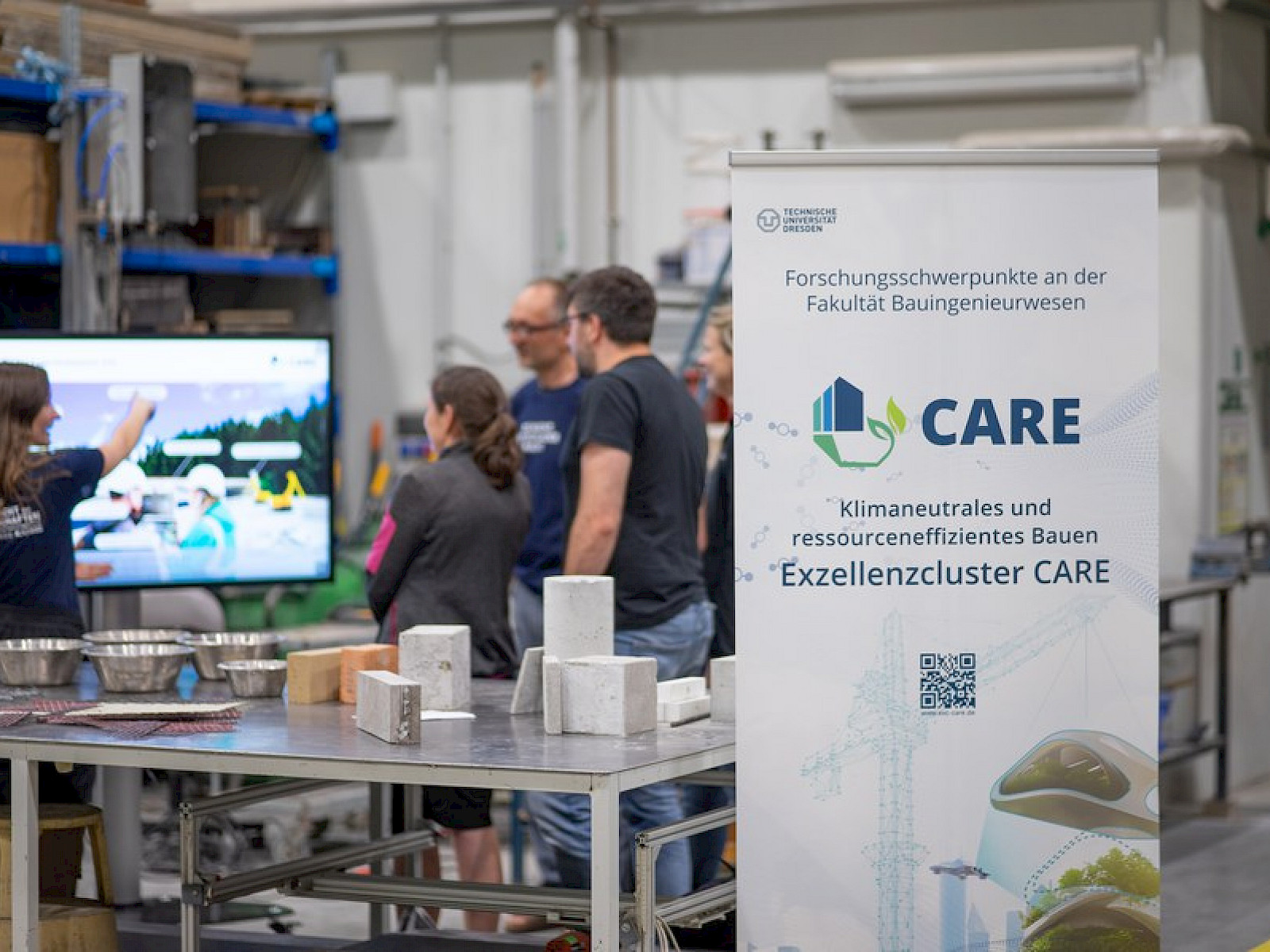 Interested people listen to a presentation by a young scientist in front of a large screen. On the table in the foreground are a few exhibits made of concrete and alternative reinforcements