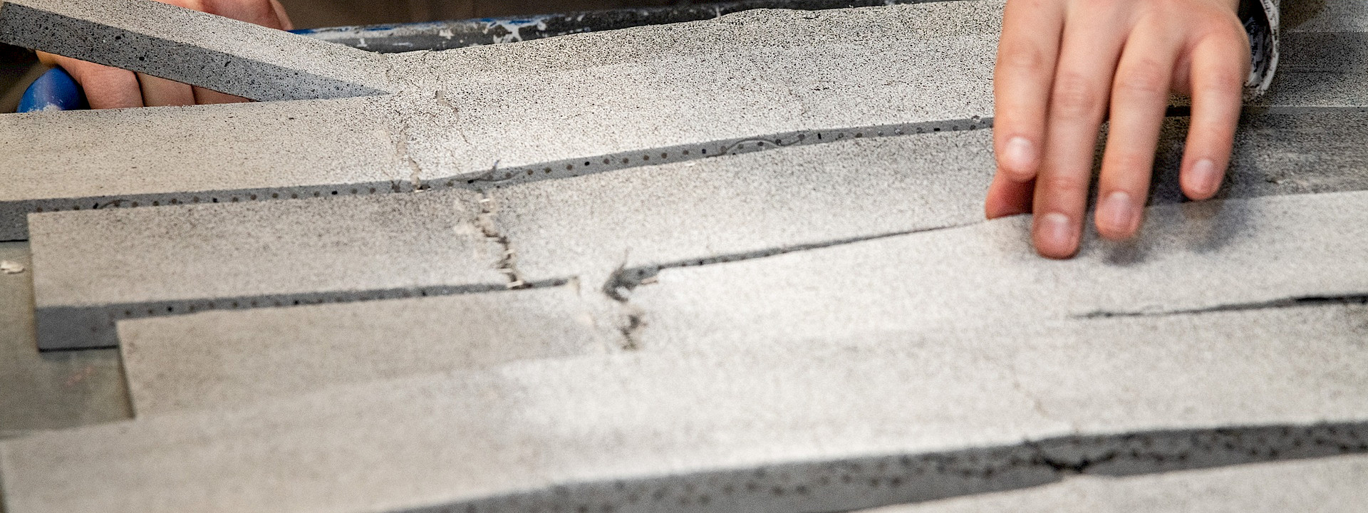 Close-up of hands inspecting and touching broken and cracked panels of grey, porous material on a workbench.