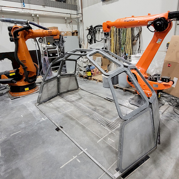 Concrete shell consisting of filled and frame modules constructed with robot-assistance. Two large, orange industrial robots in a factory hall, positioned to the left and right of an arched, grey segmented frame structure on a concrete floor.